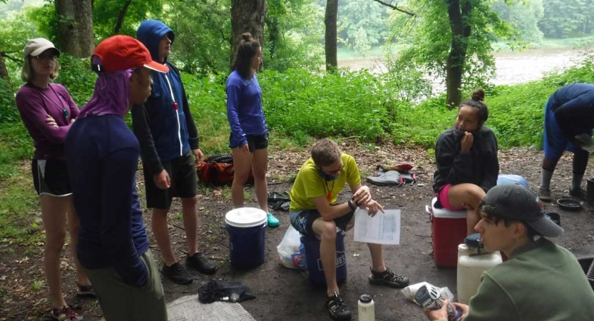 A group of people sit and stand in a wooded area with gear scattered on the ground. 